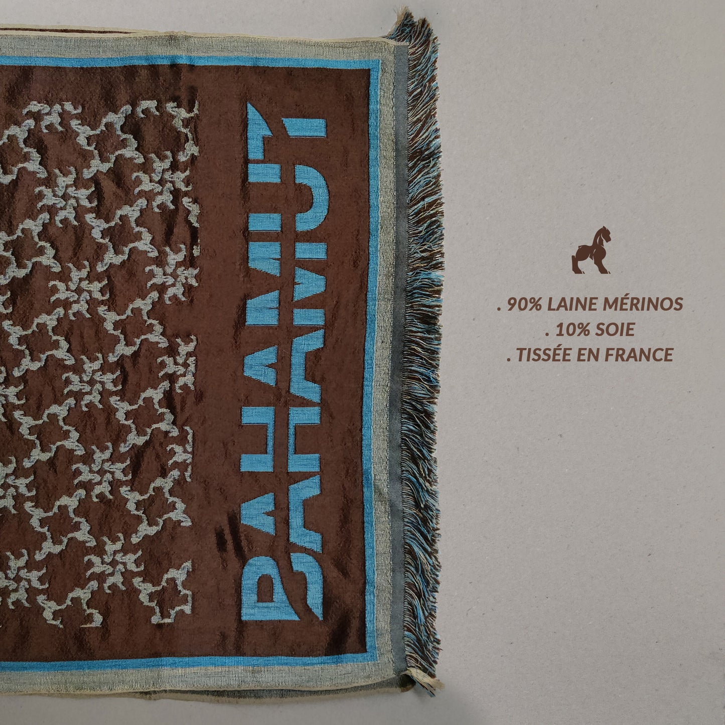 Photoshoot de l'étole pliée sur un fond neutre. on voit le monogramme et le nom de Bahamut en bleu turquoise.